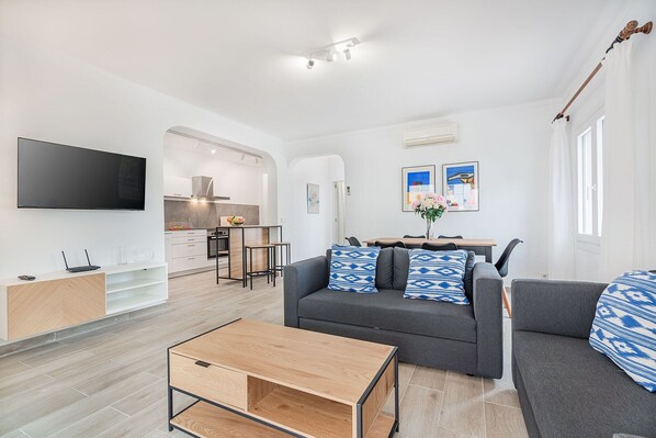Ein modernes Wohnzimmer mit einer Couch, einem Couchtisch und einem Fernseher. Es gibt eine Küchenzeile mit einem Herd und Schränken.