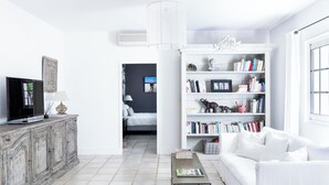 A modern living room with a white sofa, a television on a wooden cabinet, a bookshelf filled with books and decorative items, and a dining table with a magazine. - L’Orangerie – chic country house with pool, and child friendly gardens (Blauzac)