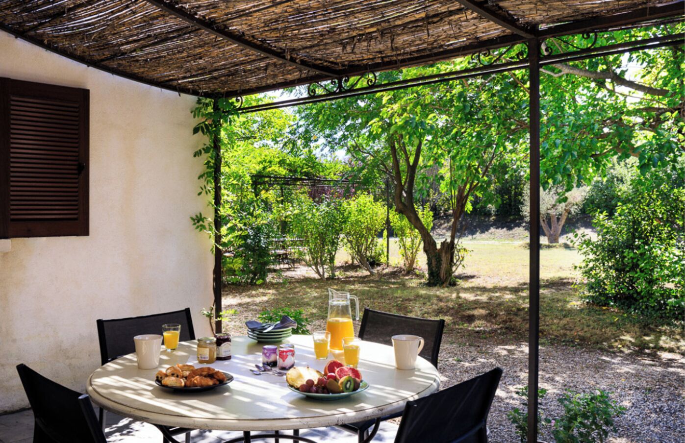 An outdoor dining area with a table set for breakfast, surrounded by chairs, and a view of a garden with trees and shrubs.