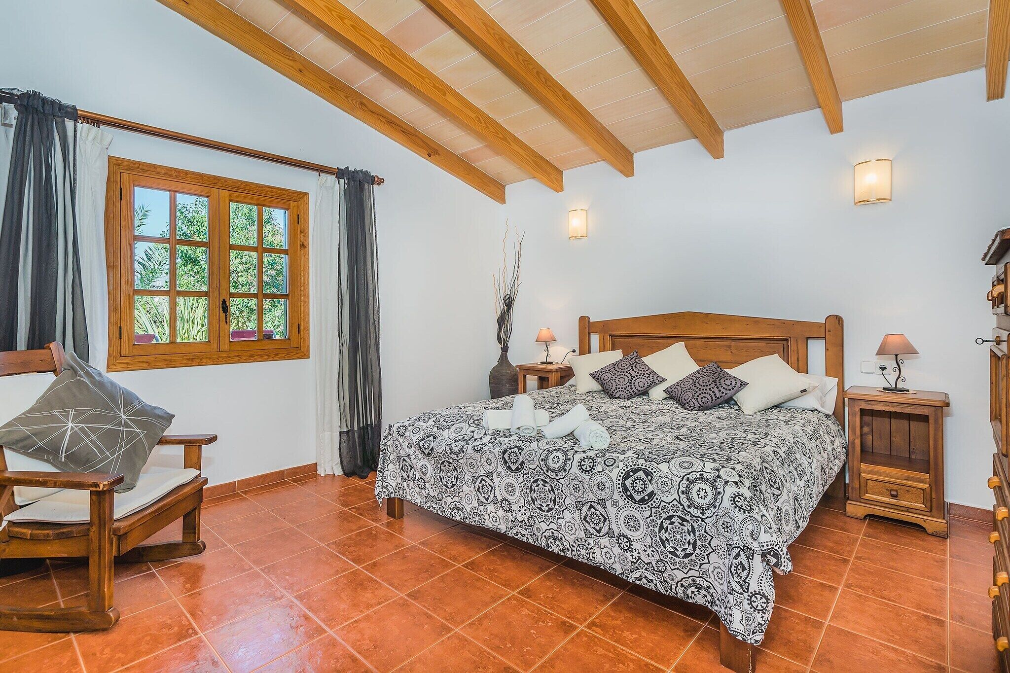A bedroom with a wooden bed, a window with curtains, a chair, a bedside table with a lamp, and a tiled floor.