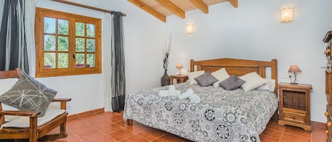 A bedroom with a wooden bed, a window with curtains, a chair, a bedside table with a lamp, and a tiled floor.