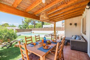 An outdoor patio with a wooden table set for breakfast, a pool in the background, and a covered seating area. - 3-Bed Villa with Pool, Puerto Pollença (Puerto Pollensa)