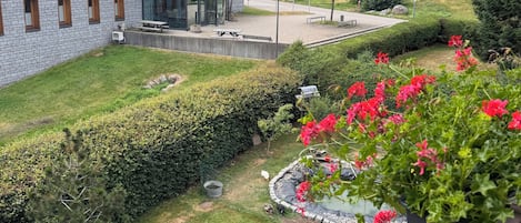 En moderne bygning med en glasfacade, en velholdt have med hække og blomster, og udsigt til en skovklædt bakke.