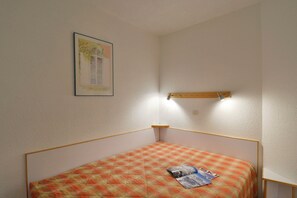 A small, single-bed room with a plaid bedspread, a framed picture on the wall, and a wooden shelf with a lamp. - travelski home classic - Résidence Themis - 2 pièces 4/5 personnes (La Plagne)