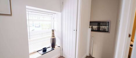A hallway with a door, a window with blinds, a vase, and a framed picture on the wall.
