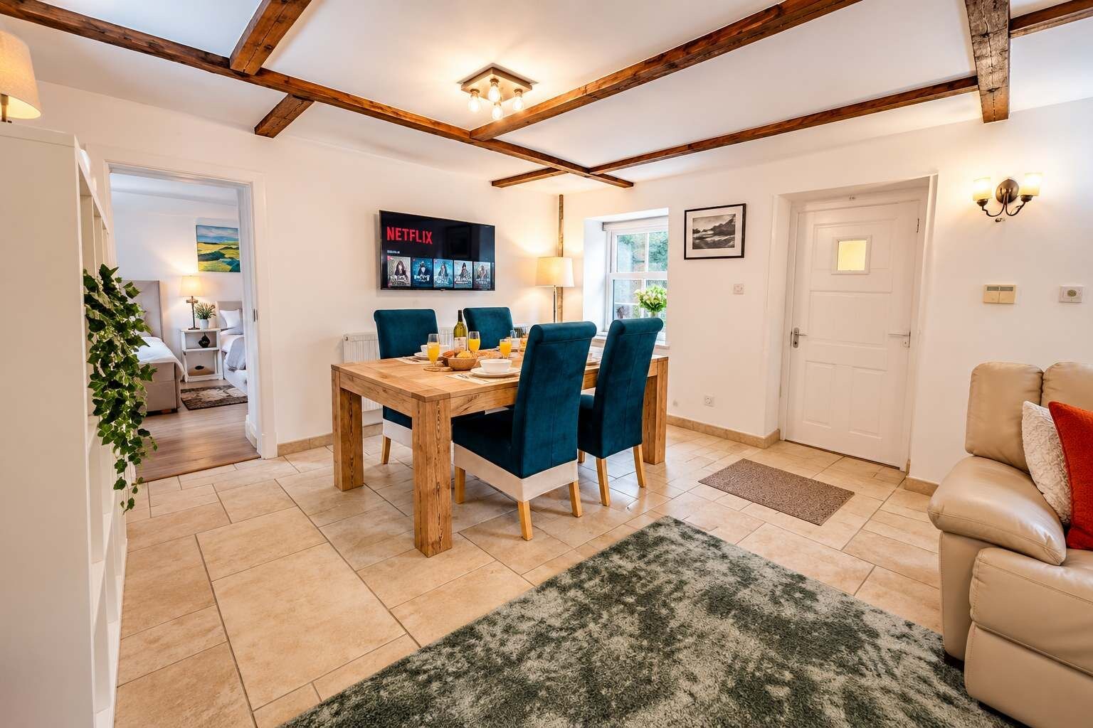 A dining area with a wooden table and blue chairs, a TV displaying Netflix, and a cozy living room with a sofa and a door.