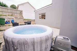 Ein Whirlpool im Freien mit Holzverkleidung, eine Terrasse mit Stühlen und ein Haus im Hintergrund.