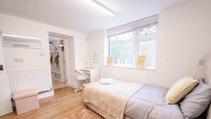 Ein Schlafzimmer mit Bett, Schreibtisch, Heizkörper, einem Fenster mit Jalousien und einer Tür zu einem anderen Raum.