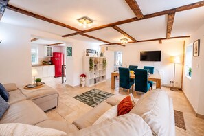A modern living room with a sofa, a dining area with chairs, a kitchen in the background, and exposed wooden beams on the ceiling.