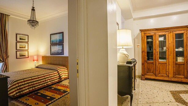 Hotellihuone, jossa on sänky, kirjoituspöytä lampun kanssa, vaatekaappi ja kattokruunu.