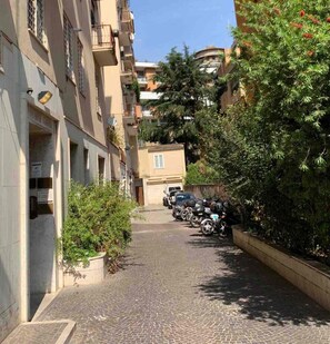 Eine enge Kopfsteinpflasterstraße, gesäumt von Wohnhäusern mit Balkonen, abgestellten Motorrädern und üppiger Bepflanzung.