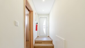 Un corridoio con una porta in legno alla fine, un estintore fissato al muro e un interruttore luminoso sulla parete.