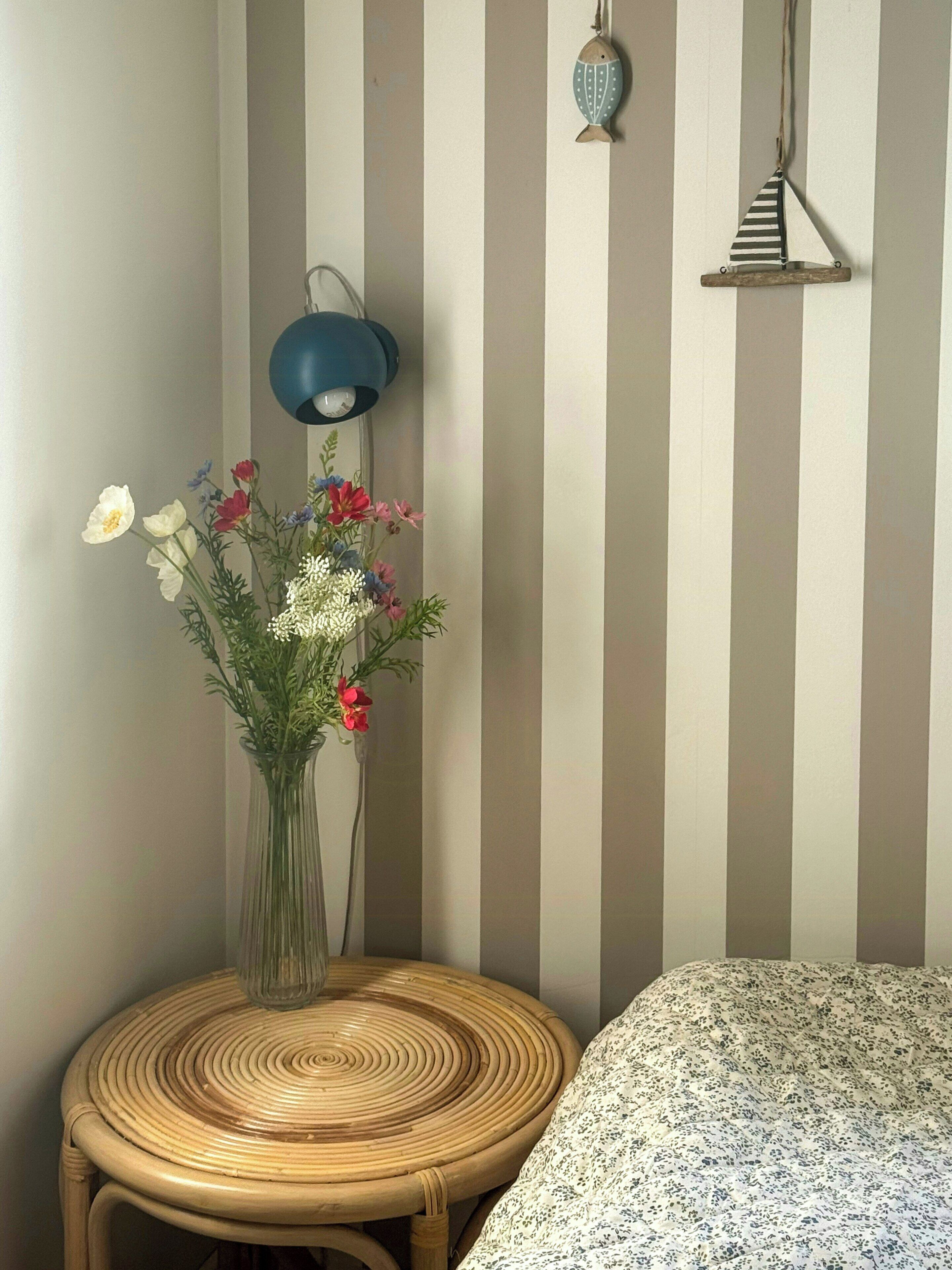 A room with a striped wall, a wicker chair, a bedside table with a lamp, and a floral bedspread.