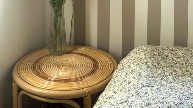 Une pièce avec un mur à rayures, une chaise en osier, une table de chevet avec une lampe et une couvre-lit à motifs floraux.