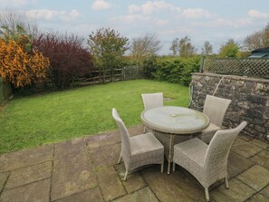 A patio with a round table and four wicker chairs, surrounded by a stone wall and a grassy lawn with trees.