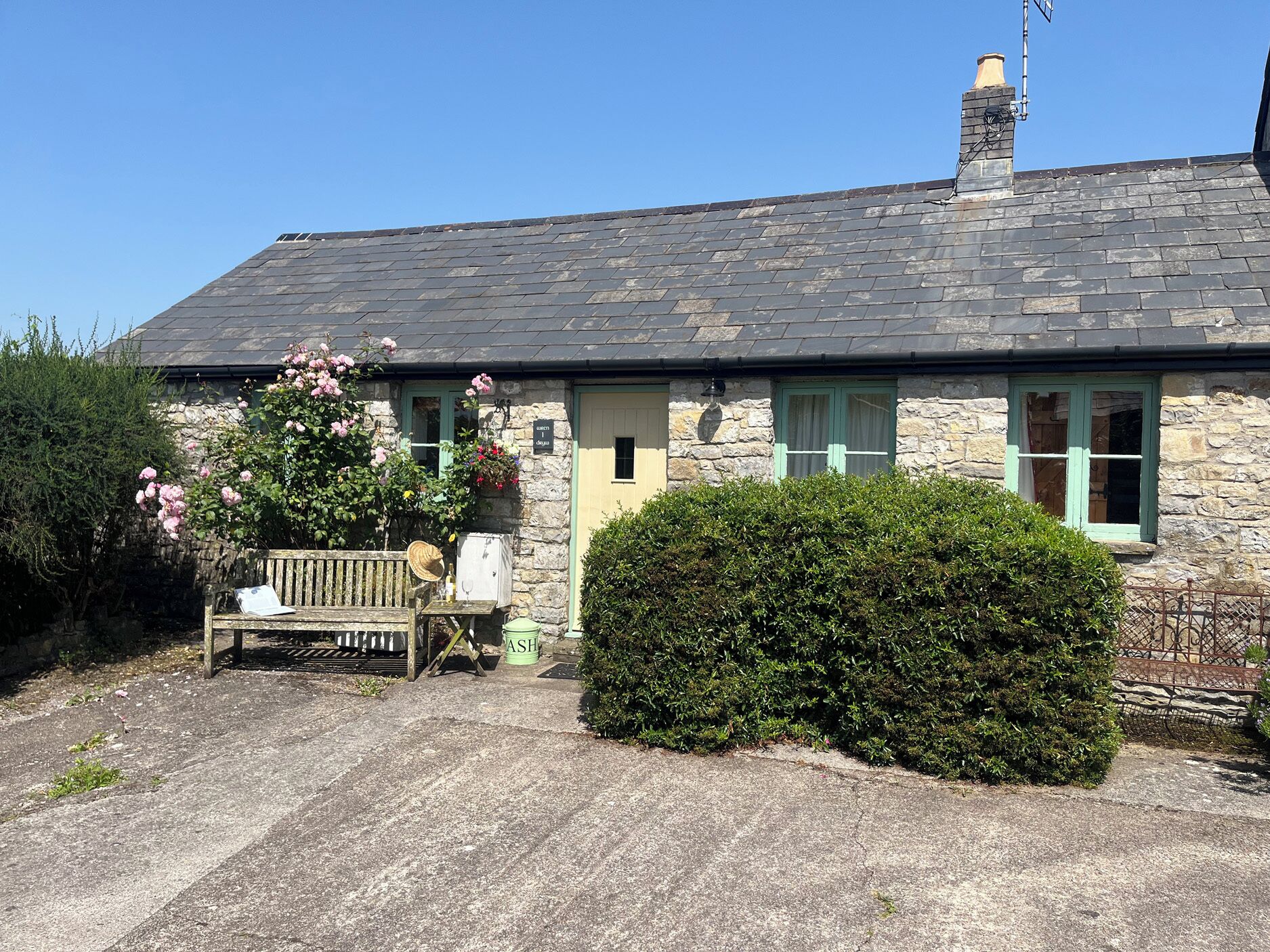 A stone house with a tiled roof, a bench, and a hedge in front.