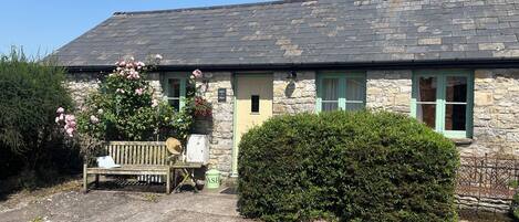A stone house with a tiled roof, a bench, and a hedge in front.