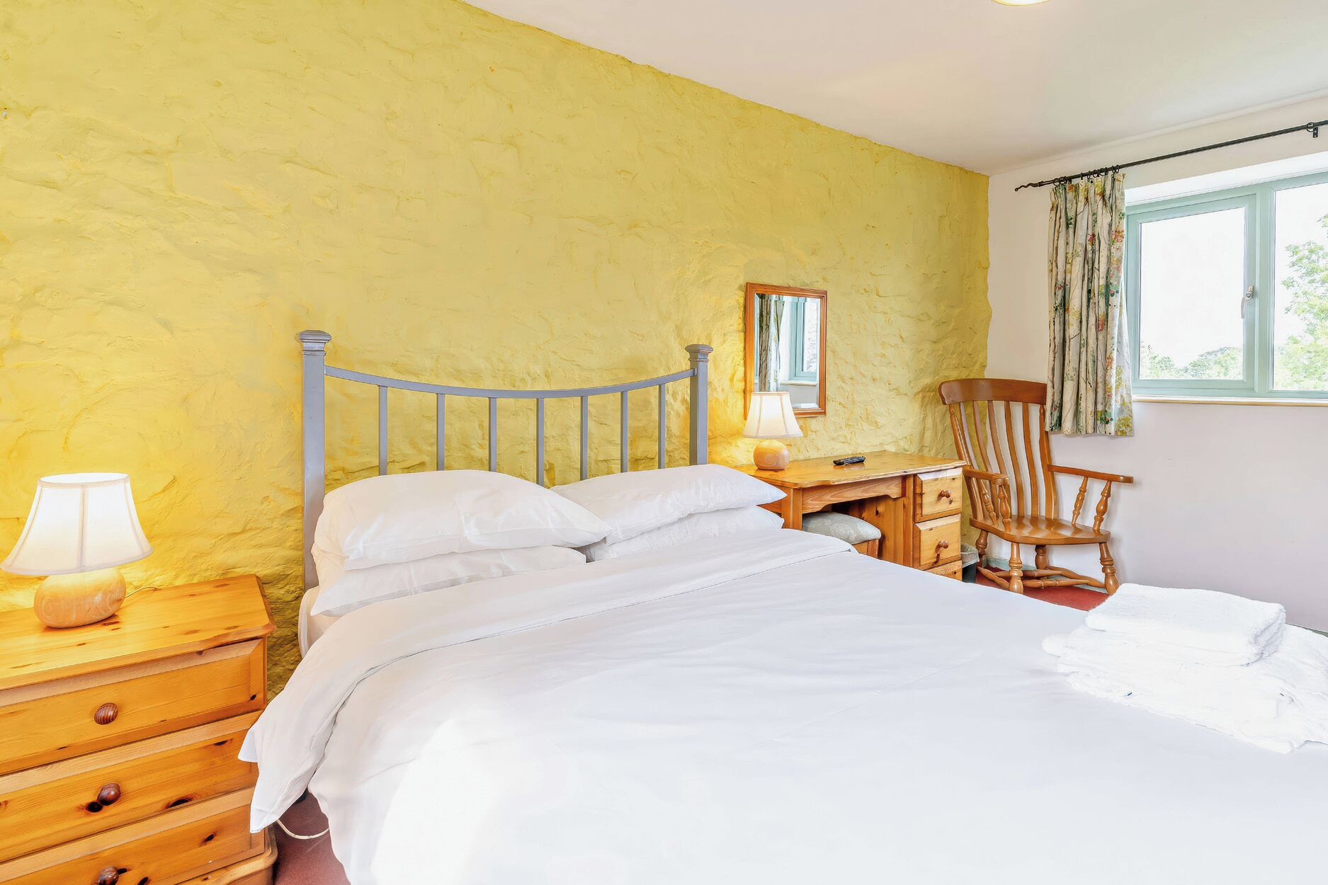 Un dormitorio con una cama, mesitas de noche de madera, una silla, una lámpara y una ventana con cortinas.