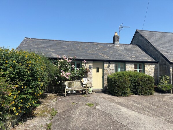 A stone building with a sloped roof, a bench, and a small garden area with flowers.