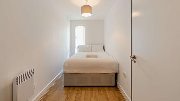 Een smalle gang die leidt naar een bed met hoofdeinde, een radiator en een deur aan de rechterkant.