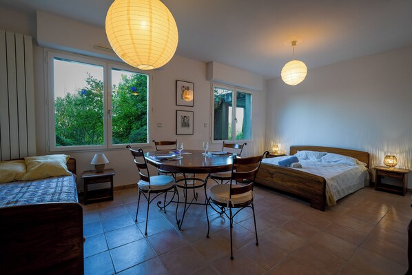 A room with a dining table, chairs, a bed, and a window with a view of greenery.