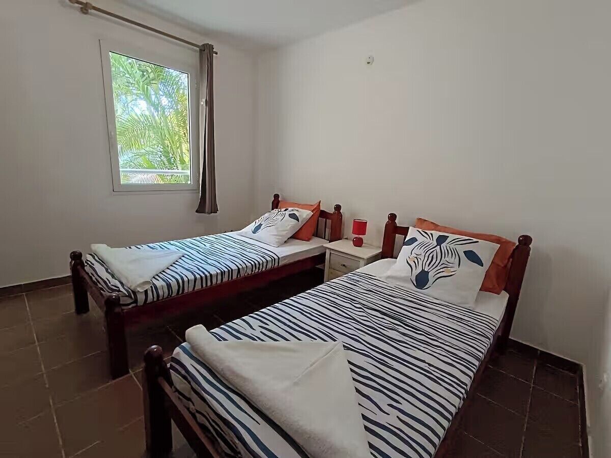 A room with two single beds, each with a wooden frame and striped bedding. There is a window with curtains, a bedside table with a lamp, and a wall-mounted light fixture.