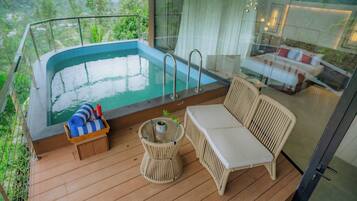 A balcony with a hot tub, wicker furniture, and a glass railing overlooking a lush landscape.