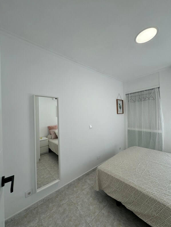A bedroom with a bed, a bedside table, a mirror, and a window with a curtain.