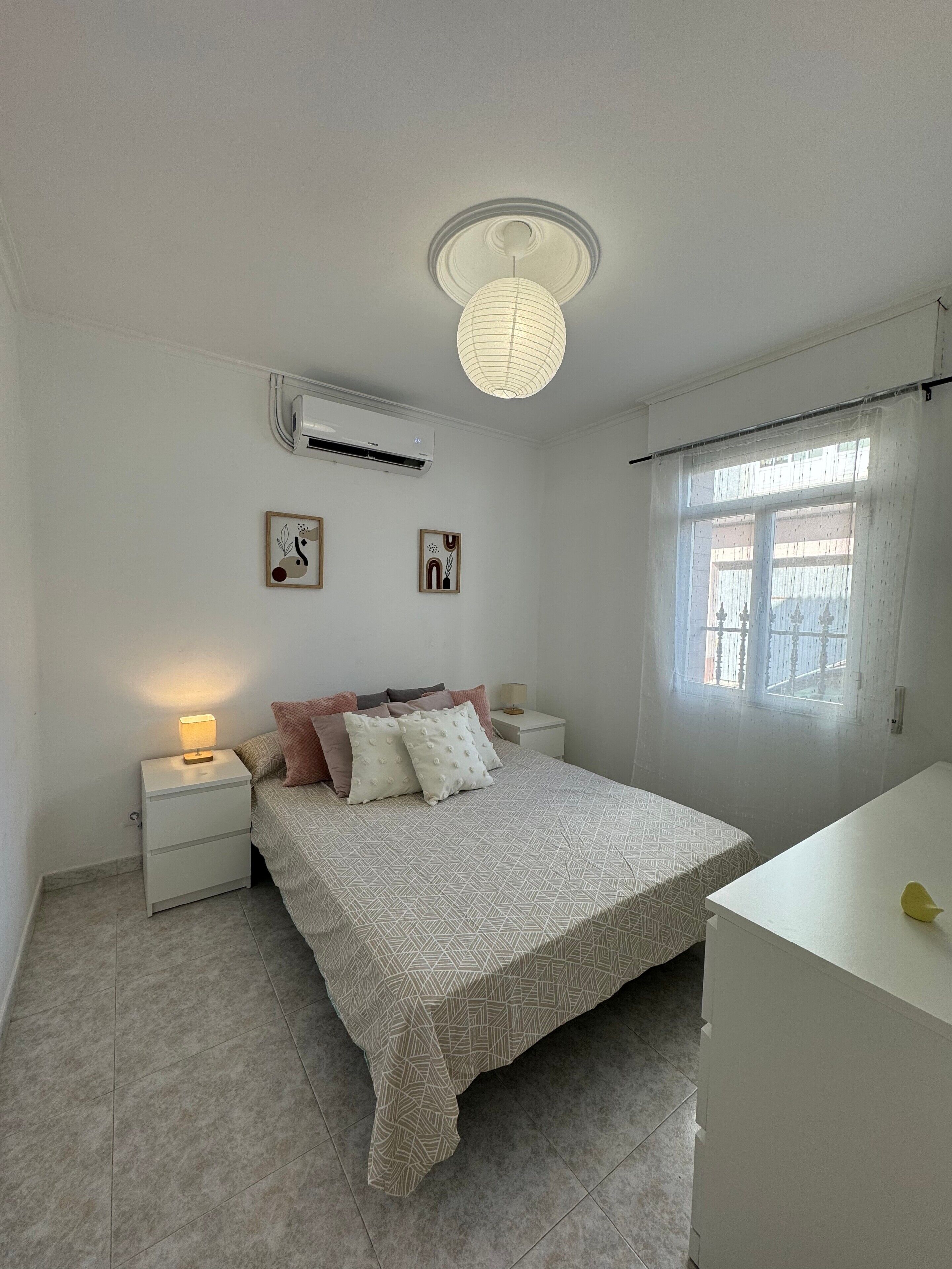 Ein Schlafzimmer mit Bett, Nachttischen, einer Nachttischlampe, einer Deckenleuchte und einem Fenster mit Vorhängen.