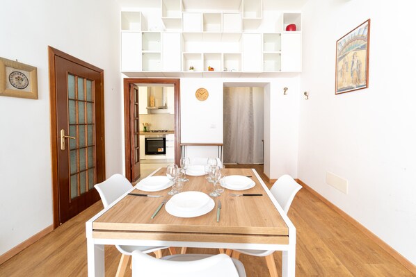 A dining area with a wooden table set for four, white chairs, and a kitchen in the background. - Appartamento in Piazzetta by Wonderful Italy (Naples)