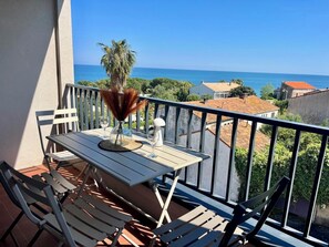 Un balcón con una mesa y sillas, un jarrón con plantas secas y vista al mar y edificios.