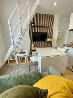 A compact living space with a staircase, a flat-screen TV mounted on a wall, a small dining area, and a kitchenette. - Cozy Duplex in the Heart of Downtown with Wi-Fi, Near the Train Station (Amiens)