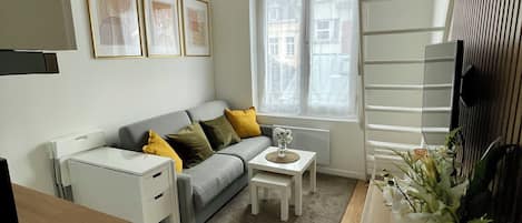 Un salón moderno con un sofá gris, una mesita blanca y una ventana con vistas a edificios.