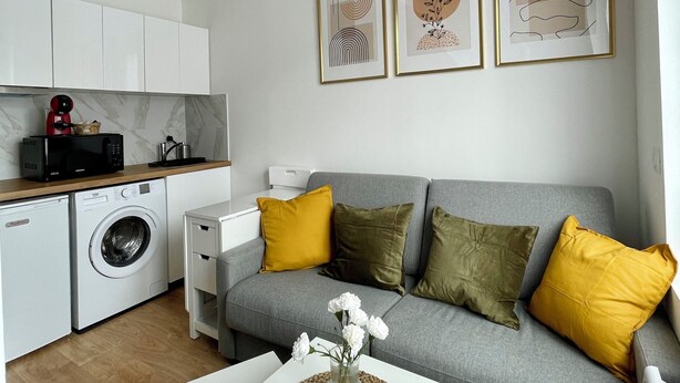 Ein moderner Wohnbereich mit grauem Sofa, gelben Kissen, einem weißen Couchtisch und einer Küchenzeile mit Geräten.