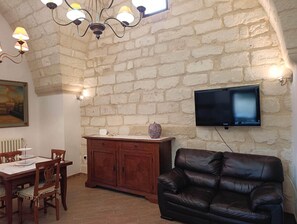 A room with a stone wall, a television mounted on it, a wooden cabinet, a leather sofa, and a dining table with chairs. - Shadows of Stone - Historic Mansion (Melendugno)