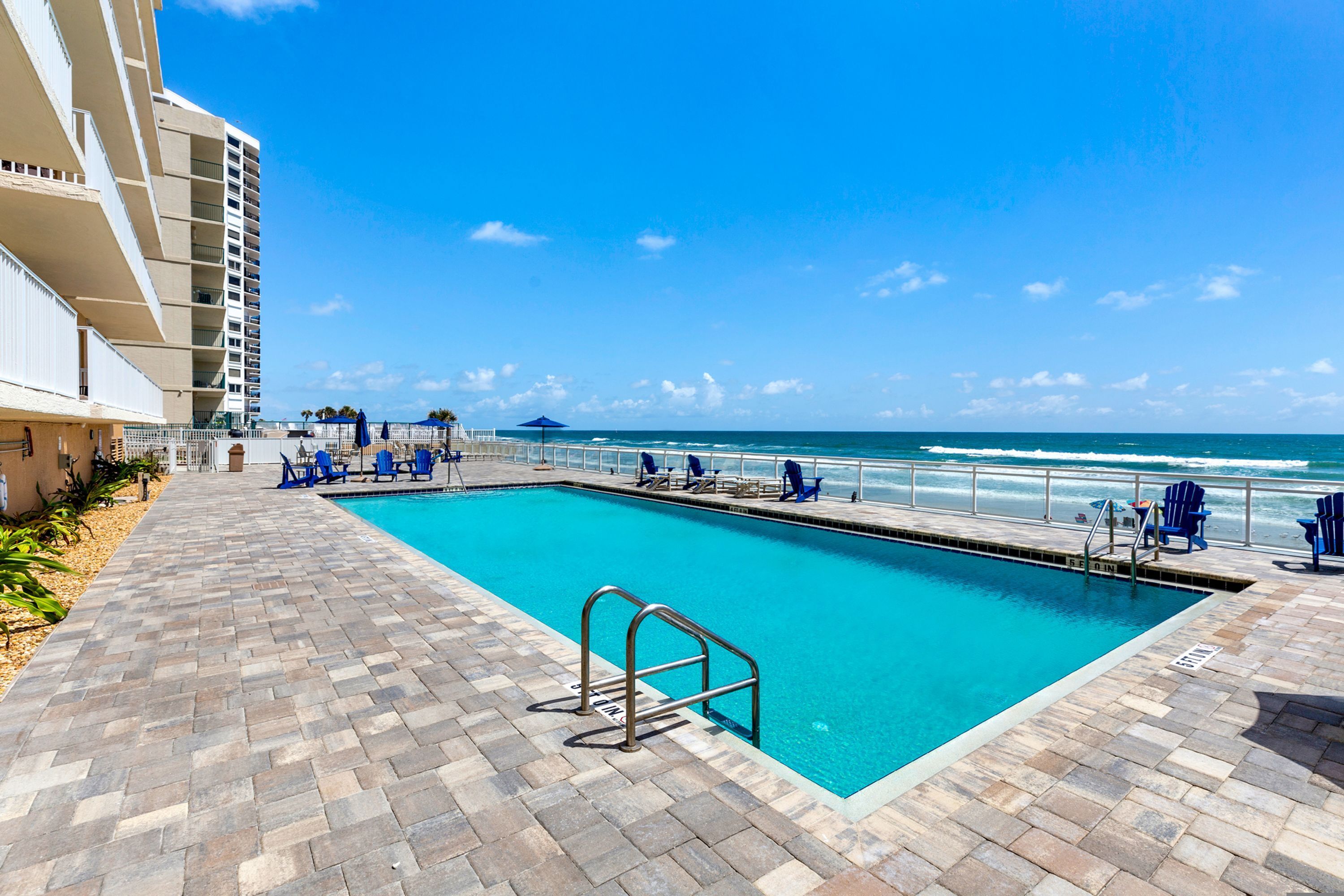 A swimming pool with a view of the ocean, a brick pathway, and blue chairs.