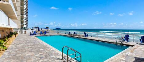 A swimming pool with a view of the ocean, a brick pathway, and blue chairs.