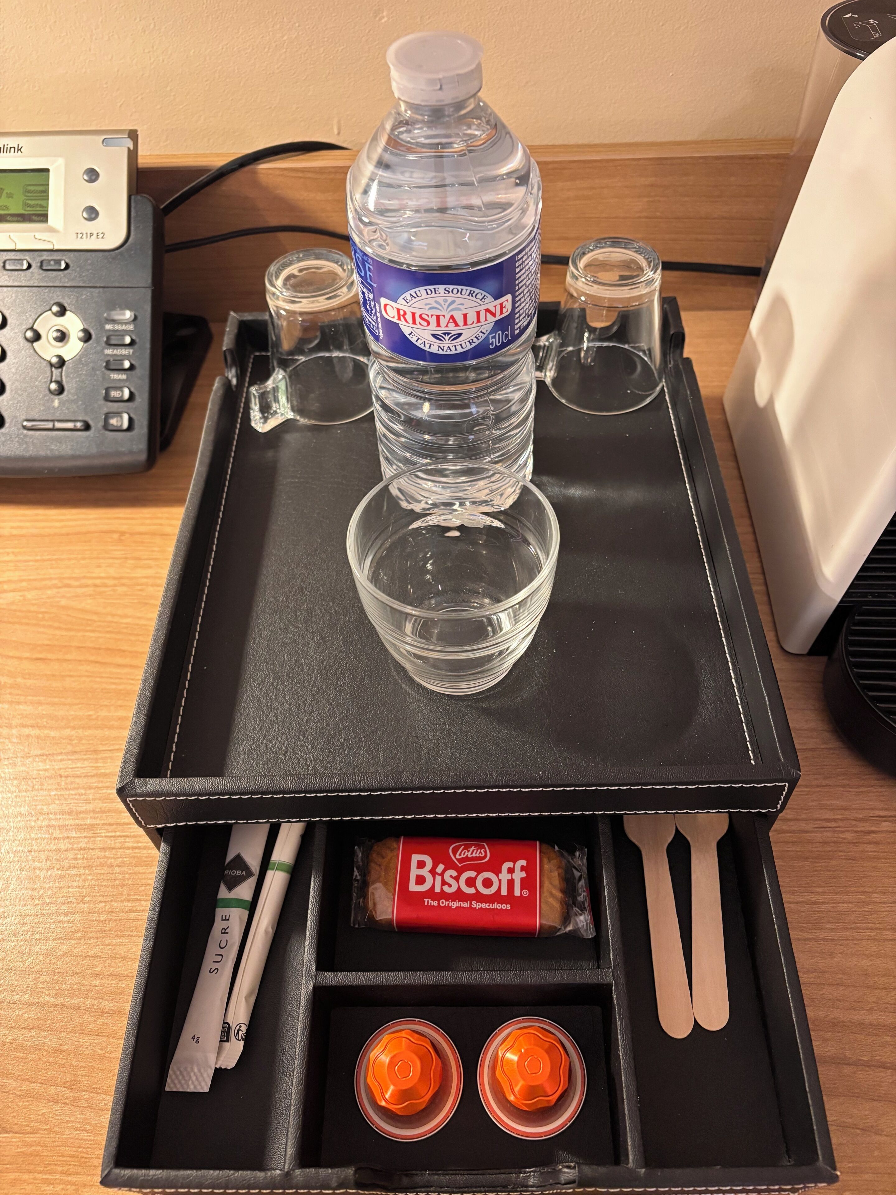 A tray with a water bottle, glasses, a box containing a biscuit packet, a spoon, and two orange capsules.