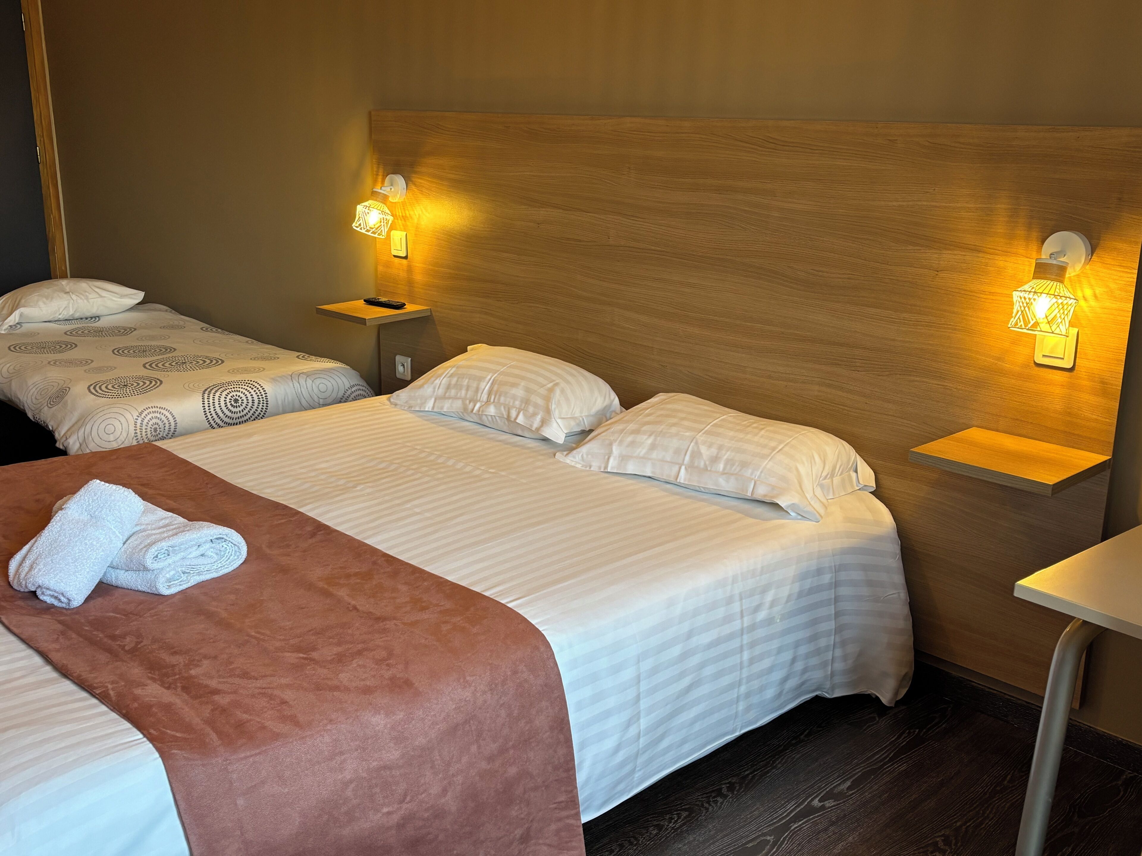 Habitación de hotel con dos camas, cabecera de madera, mesitas de noche con lámparas y una toalla doblada con cuidado sobre la cama.