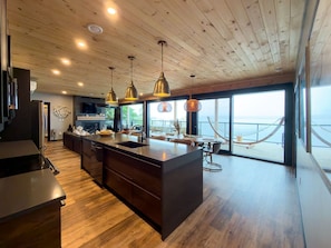 A modern kitchen with wooden flooring, a central island, pendant lighting, and a view of the sea through large windows.