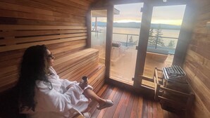 Innenansicht einer Holzhütte mit einer Bank, einer Person im Bademantel und einem großen Fenster mit Blick auf ein Gewässer.