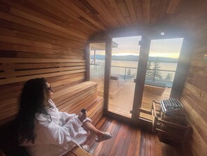 Interior de una cabaña de madera con un banco, una persona con bata y un ventanal con vistas a un cuerpo de agua.