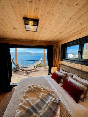 A wooden cabin bedroom with a large bed, a hammock, and a view of the lake.