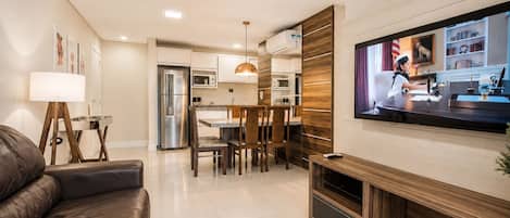 A modern living room with a leather sofa, a dining area with wooden chairs, a kitchen with white cabinets, and a TV mounted on the wall.
