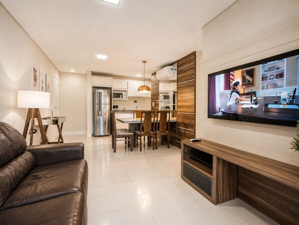 A modern living room with a leather sofa, a dining area with wooden chairs, a kitchen with white cabinets, and a TV mounted on the wall. - Porto Fino by LikeHome  (Bombinhas)