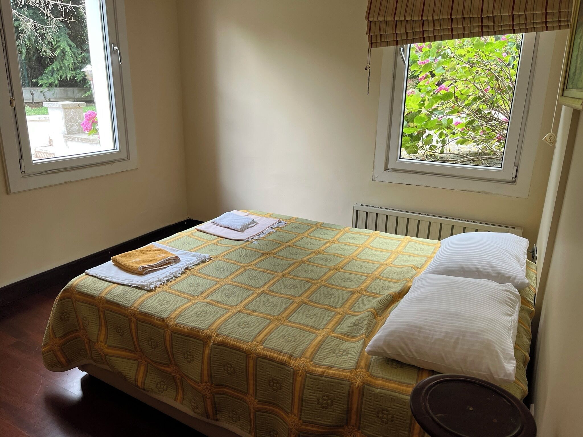 A single bed with a patterned bedspread, two pillows, a folded towel, and a bedside table with a lamp.