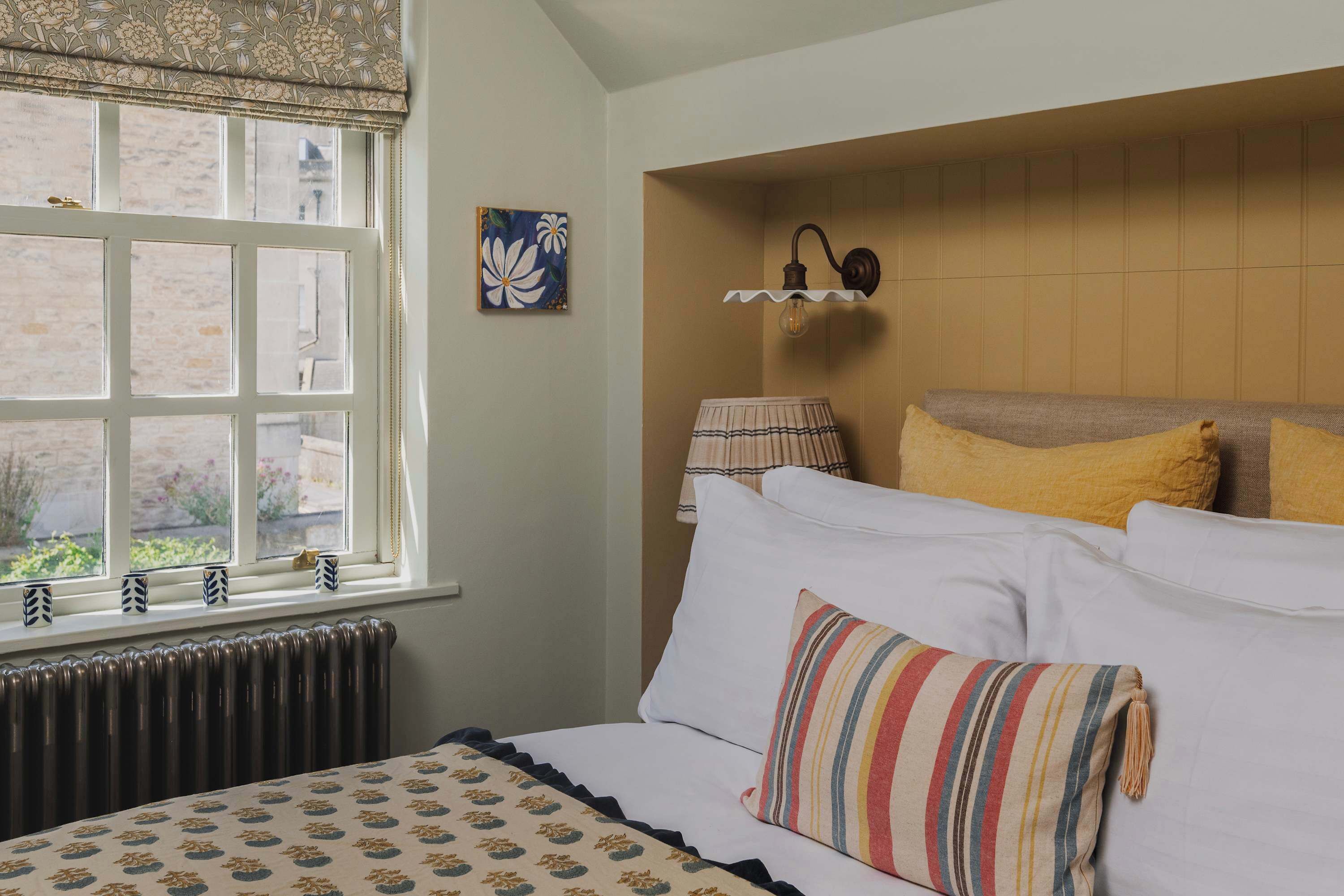 Une chambre avec une grande fenêtre, un radiateur, un lit avec une literie blanche et des oreillers à rayures, une lampe fixée au mur et un tableau.