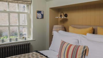 Une chambre avec une grande fenêtre, un radiateur, un lit avec une literie blanche et des oreillers à rayures, une lampe fixée au mur et un tableau.
