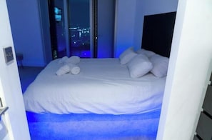 A hotel room with a bed, a view of the city at night, and a blue carpet. - Luxury 2-Bed Penthouse | Stunning City Views | Heart of Manchester (Manchester)