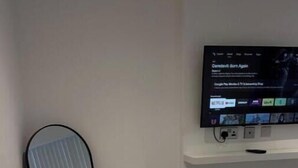 Een kamer met een flatscreen televisie aan de muur, een staand spiegel en een wit schap onder de televisie.